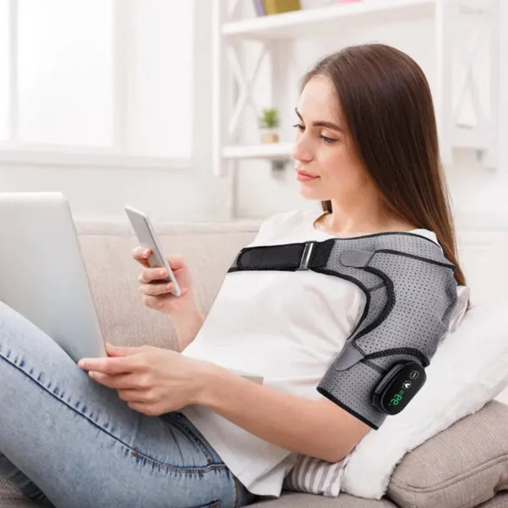  Woman relaxing at home using the RelaxRiser™ HeatFlex Shoulder Wrap while working on a laptop and phone, showcasing its hands-free, ergonomic design for daily comfort and wellness.